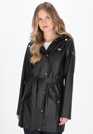 Nuori nainen musta vyöllä varustettu sadetakki, jossa on huppu ja painonapit, seisoo valkoista taustaa vasten.