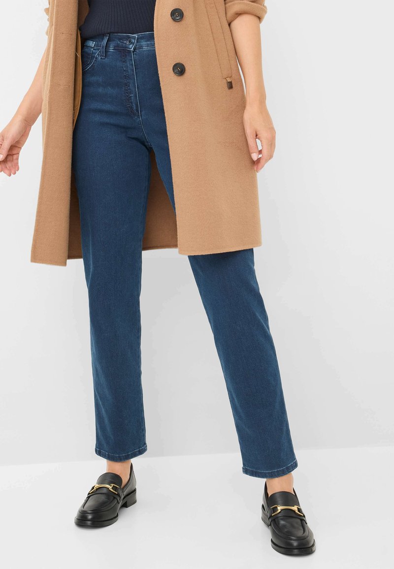 Jeans droits bleu marine avec une légère décoloration, associés à un manteau en laine beige et des mocassins en cuir noir avec un accent doré.