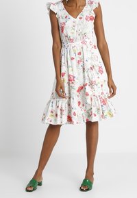 Blumenmusterkleid mit weißem Hintergrund, Rüschenärmeln, V-Ausschnitt, elastischem Bund und einem gestuften Rüschensaum. Getragen mit grünen Sandalen.