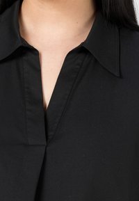Chemise noire à col, avec un décolleté en V profond, fabriquée en tissu lisse, avec des lignes épurées et des détails de couture subtils.