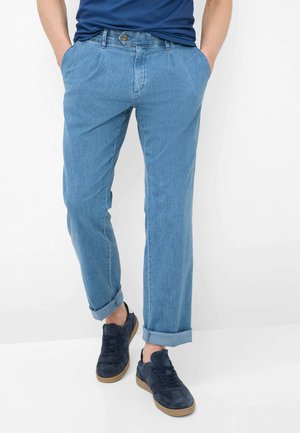 Jeans azzurri chiari slim fit con risvolti, indossati con sneakers blu navy e una camicia blu scuro infilata, mani nelle tasche.