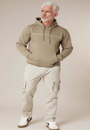 Homme d'âge moyen aux cheveux gris et à la barbe, portant un sweat à capuche beige avec le texte « DEELUXE ORIGINAL », un pantalon cargo crème et des baskets blanches, debout.