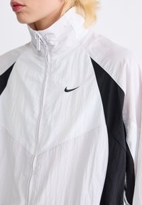 Veste zippée blanche et noire avec un col haut, en matériau léger, et un logo Nike sur la poitrine. Présente des blocs de couleur contrastants.