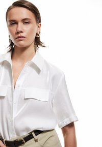 White short-sleeve blouse in glossy fabric, featuring a collar and two chest pockets. Paired with a beige belt and pants.