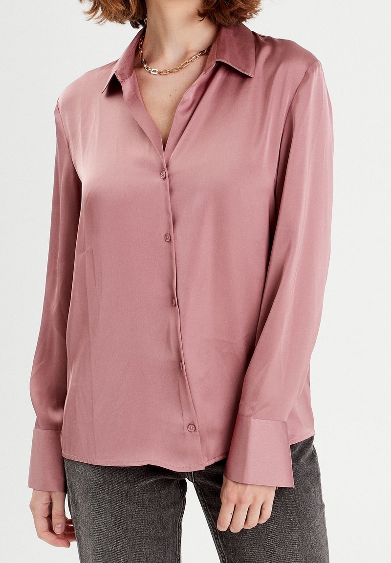 Femme portant un chemisier en satin rose poudré boutonné avec de longues poignets et un jean gris foncé, accessoirisée d'un collier chaîne en or.