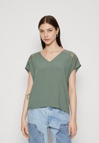 Top verde oliva a V con maniche corte, con dettagli in pizzo sulle spalle e una texture morbida e liscia. Abbinato a jeans leggeri blu chiaro a fantasia.
