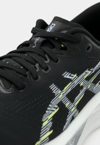 Chaussure de sport noire avec un dessus texturé, des motifs dynamiques gris et jaune fluo, et un design traditionnel à lacets.