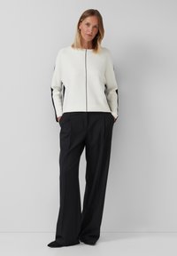 Vrouw staand met haar handen in de zakken, gekleed in een witte trui met zwarte strepen en een wijdvallende zwarte broek, tegen een lichtgrijze achtergrond.