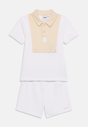 Balta īsroku polo krekls ar bēša apkakles pārklājumu un atbilstošiem baltiem šortiem, uz šortiem ir mazs "BOSS" logotips.