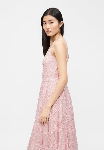 Νεαρή γυναίκα με μακριά σκούρα μαλλιά φοράει μαν sleeveless ροζ φόρεμα με δαντέλα και λουλούδια, στέκεται πλαγίως μπροστά σε απλό λευκό φόντο.