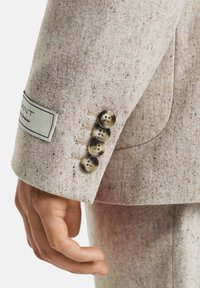 Nahaufnahme eines beigen, gesprenkelten Blazers mit vier braunen Knöpfen und einem sichtbaren Markenetikett, getragen von einer Person, die ihre Hand entspannt hält.