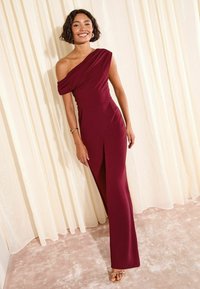 Rochie bordeaux, cu o singură umăr, dintr-un material fin, cu detalii drapate și o fanta înaltă până la coapsă, asortată cu sandale aurii.