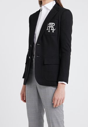 Vrouw draagt een zwart colbert met een wit geborduurd logo op de zak, een wit overhemd met knoopjes en grijze geruite broek, staande tegen een witte achtergrond.