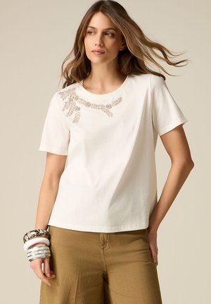Woman with light brown hair wears white short-sleeve shirt with beaded floral detail and khaki pants, accessorized with chunky bracelets.