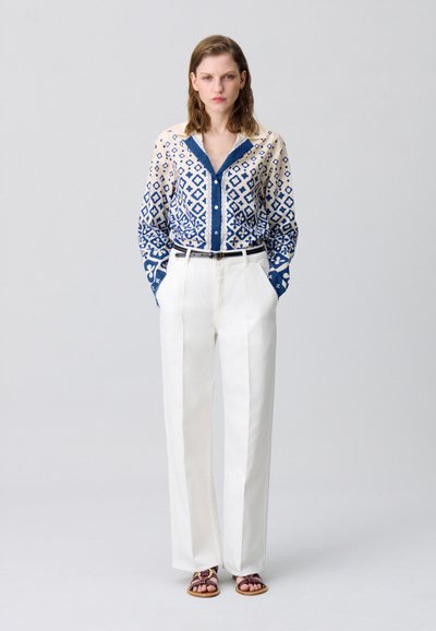 Camicia a maniche lunghe con un motivo geometrico blu e crema, abbinata a pantaloni bianchi a vita alta e sandali bassi.
