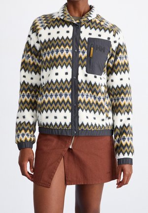 Veste en laine avec motifs zigzag jaunes, noirs et gris, poche poitrine grise et boutons, portée avec une jupe marron avec une fente zippée à l'avant.