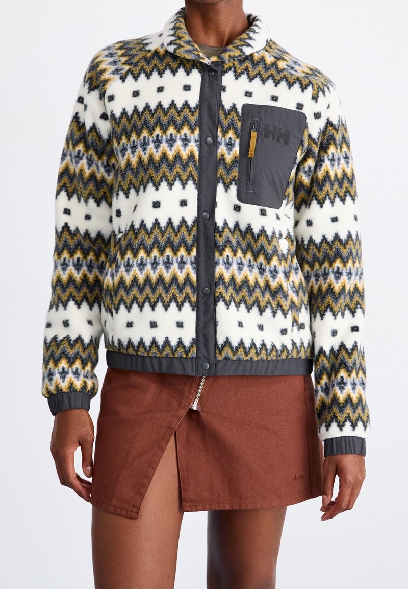 Veste en laine avec motifs zigzag jaunes, noirs et gris, poche poitrine grise et boutons, portée avec une jupe marron avec une fente zippée à l'avant.