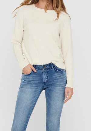 Pull tricoté blanc cassé avec un col rond, poignets côtelés, porté avec un jean denim bleu comprenant des poches et une légère décoloration.