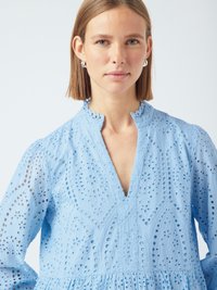 Γυναίκα με ίσια μαλλιά μέχρι τους ώμους, φορώντας ασημένια σκουλαρίκια κρίκους και μια ανοιχτό μπλε μπλούζα με κεντητές λεπτομέρειες και γιακά σε σχήμα V.