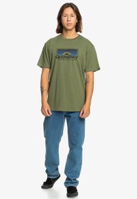 Olive green cotton t-shirt with Quiksilver logo in blue and black, paired with light blue denim jeans and black shoes, standing posture.