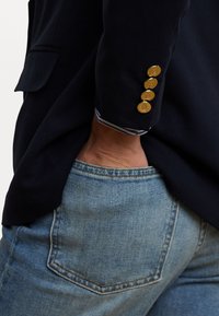 Marineblazer met gouden decoratieve knopen op de manchetten. Blauw-witte gestreepte voering zichtbaar. Draagt over lichtblauwe denim jeans.