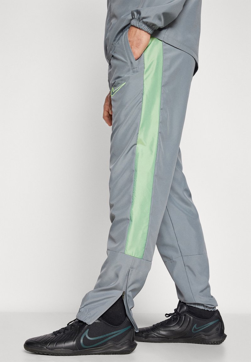 Pantalons de jogging gris avec une bande latérale verte, des manches zippées et un petit logo Nike noir sur la cuisse gauche ; fabriqués en matériau lisse et léger.