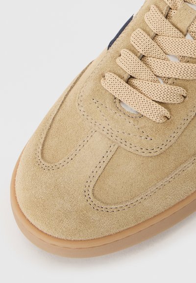 Baskets en daim beige avec coutures renforcées, bout rond et lacets tissés beige. Semelle en caoutchouc de couleur brun clair. Accentué par des détails subtils.