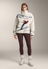 Biely fleecový pulóver s lyžiarskym motívom, vysokým golierom a polovičným zipsom. Kombinovaný s vínovo červenými legínami a bielymi zimnými topánkami. Mäkká textúra.