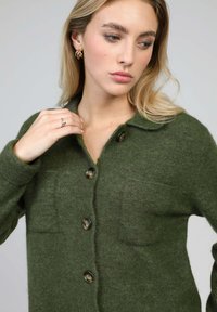 Cardigan vert en matière douce, doté d'un col, de poches avant et de boutons foncés. Tricot texturé avec une coupe décontractée.