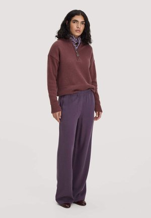Frau mit lockigem dunklem Haar, die einen mauvefarbenen Strickpullover, violette weite Hosen und einen gemusterten Schal um den Hals trägt und vor einem schlichten Hintergrund steht.