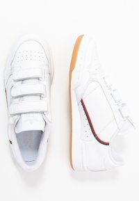 Vita sneakers med tre kardborreband, röda och gröna sidopaneler, perforerade detaljer och en ljusbrun gummisula.