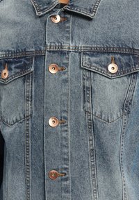 Jeansowa kurtka z jasnoniebieskiego materiału, klasycznym kołnierzem, dwoma kieszeniami na piersi oraz mosiężnymi guzikami z metalicznym wykończeniem i detalami przeszycia.