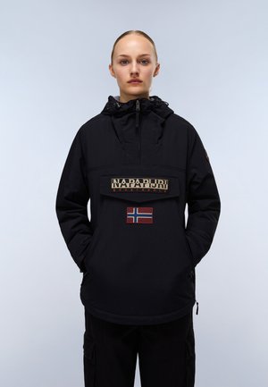 Nainen katsoo eteenpäin, päällään musta Napapijrin vetoketjullinen takki, kädet taskuissa, ja takissa on etupuolella Norjan lipun merkki.