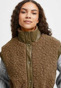 Vest de fleece texturat, de culoare maronie, cu guler matlasat de culoare verde măsliniu și nailon cu capse, având un amestec de materiale moi și netede.