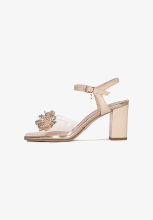 Beżowy sandał na szerokim obcasie z paskiem wokół kostki, przezroczystym paskiem z przodu i ozdobą w postaci przezroczystego kwiata przy palcach.