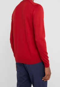 Czerwony sweter dzianinowy z długimi rękawami i okrągłym dekoltem, gładka tekstura, prążkowane mankiety i dół, noszony na granatowych spodniach.