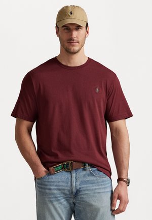 Bordeauxrotes Baumwoll-T-Shirt mit Rundhalsausschnitt und kurzen Ärmeln, versehen mit einem kleinen grünen Logo auf der linken Brust. Kombiniert mit blauen Jeans und einem strukturierten Gürtel.