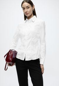 Λευκό πουκάμισο με μακριά μανίκια και κουμπιά, με μυτερό γιακά, συνδυασμένο με μαύρο παντελόνι. Κρατάει μια μικρή δερμάτινη τσάντα μπορντό.