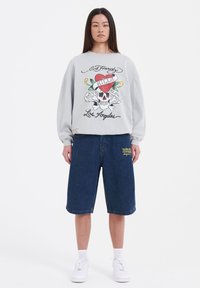 Grå sweatshirt med grafisk tryck av ett skalle och hjärta, i kombination med knälånga denimshorts. Vita sneakers fulländar outfiten.