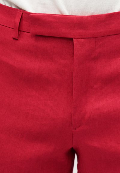 Κοντινή λήψη της μέσης κόκκινων ραμμένων παντελονιών με ζώνη μέσης, θηλάκια για ζώνη και εμπρόσθιο κλείσιμο φερμουάρ σε λευκό φόντο.