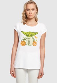 T-shirt bianca con un personaggio dei cartoni animati verde che tiene dei secchi di caramelle a forma di zucca arancione, con un design di stelle e un pipistrello. Maniche corte, vestibilità comoda.
