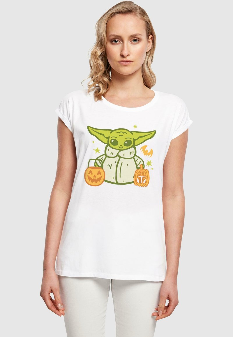 T-shirt bianca con un personaggio dei cartoni animati verde che tiene dei secchi di caramelle a forma di zucca arancione, con un design di stelle e un pipistrello. Maniche corte, vestibilità comoda.