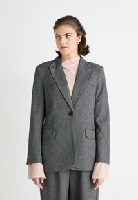 Grijze op maat gemaakte blazer met notch lapels, een enkele knoopsluiting en voorzakken. Heeft opvallende, gestructureerde roze manchetten die onder de mouwen uit komen.