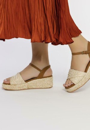 Sandalias de cuña con parte superior de yute y paja trenzados, correas en tono marrón claro y suela de corcho. El color marrón claro se combina con una falda de cintura plisada naranja.
