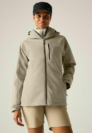 Femme portant une veste à capuche beige, un short beige et une casquette noire, debout avec une main dans la poche devant un fond uni.