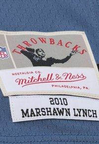 Das Mitchell & Ness-Label hat einen grauen Hintergrund mit rotem Text, auf dem "THROWBACKS" steht, und einer schwarzen Silhouette, darunter steht "MARSHawn LYNCH 2010."