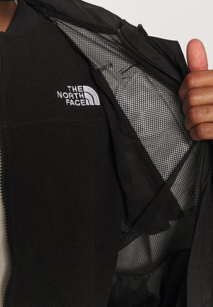 La main d'une personne ouvre une veste noire pour montrer la couche intérieure en polaire noire avec le logo blanc "The North Face" sur la poitrine.