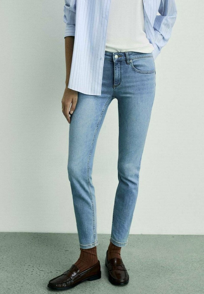 Personne portant un jean skinny bleu clair, des chaussettes marron côtelées, des mocassins marron foncé, un haut blanc et une chemise rayée bleu clair, debout à l'intérieur.