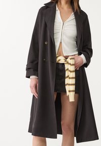 Schwarzer Trenchcoat mit großen Revers, doppelreihigem Design und umgeschlagenen Ärmeln, getragen über einem weißen Reißverschluss-Oberteil und einem schwarzen Jeansrock.