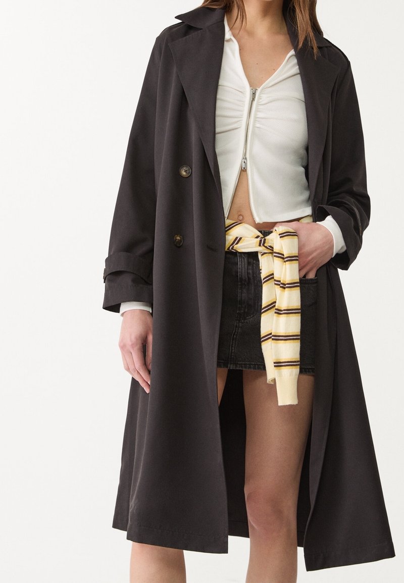 Schwarzer Trenchcoat mit großen Revers, doppelreihigem Design und umgeschlagenen Ärmeln, getragen über einem weißen Reißverschluss-Oberteil und einem schwarzen Jeansrock.